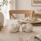 Two white ceramic cat-shaped salt and pepper shakers resting on a beige table runner in a styled dining room setting.