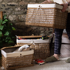 Three woven wicker storage baskets with white fabric liners are shown in a home setting. A person is holding the largest basket, while two smaller baskets sit on a rug in front of a rustic stone wall.