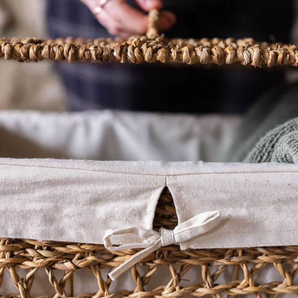 Close-up of a woven natural-fiber storage basket with a light beige fabric liner and tie, shown with a person's hands holding the handle in the background.
