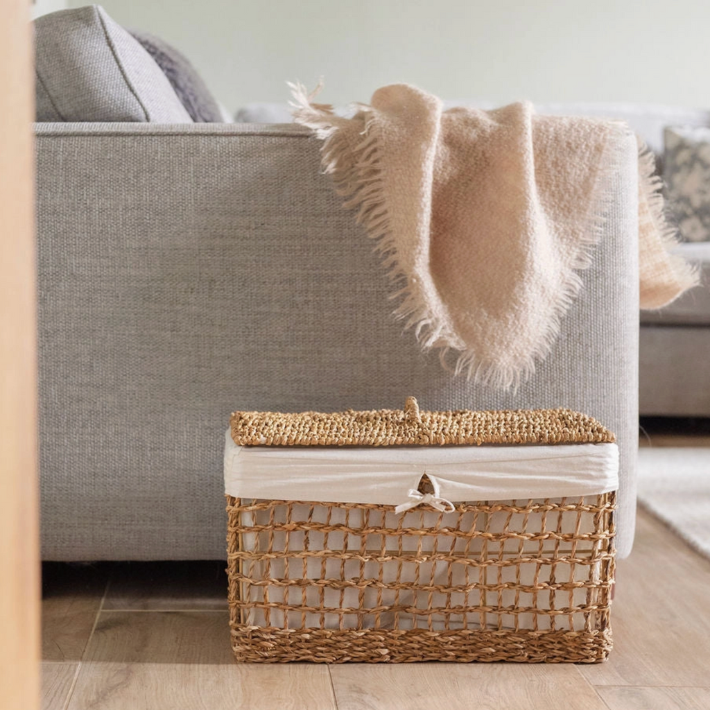A woven rectangular storage basket with a lid and white fabric liner sits on a wood floor in front of a light gray sofa with a beige throw blanket draped over it.