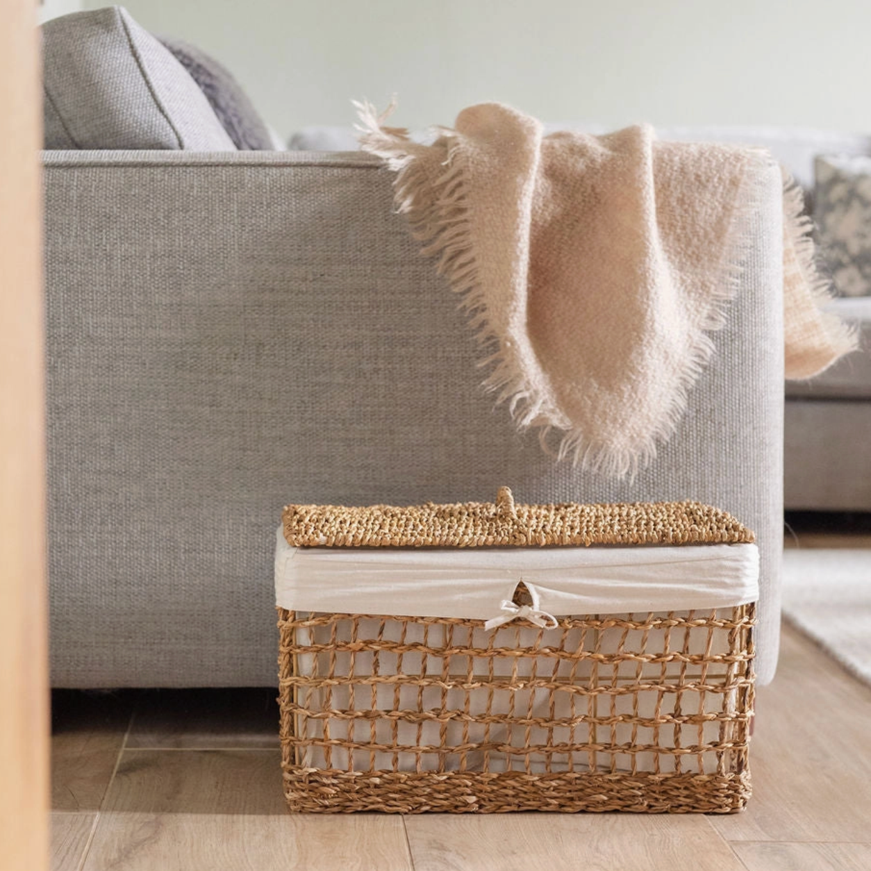 A woven rectangular storage basket with a lid and white fabric liner sits on a wood floor in front of a light gray sofa with a beige throw blanket draped over it.
