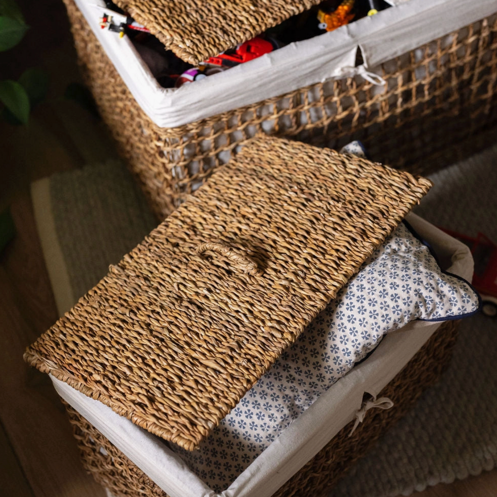 A woven natural-fiber storage basket with a fitted white fabric liner and a matching lid, shown with a patterned pillow inside. Another basket is partially visible in the background on a wooden floor.