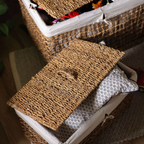 A woven natural-fiber storage basket with a fitted white fabric liner and a matching lid, shown with a patterned pillow inside. Another basket is partially visible in the background on a wooden floor.