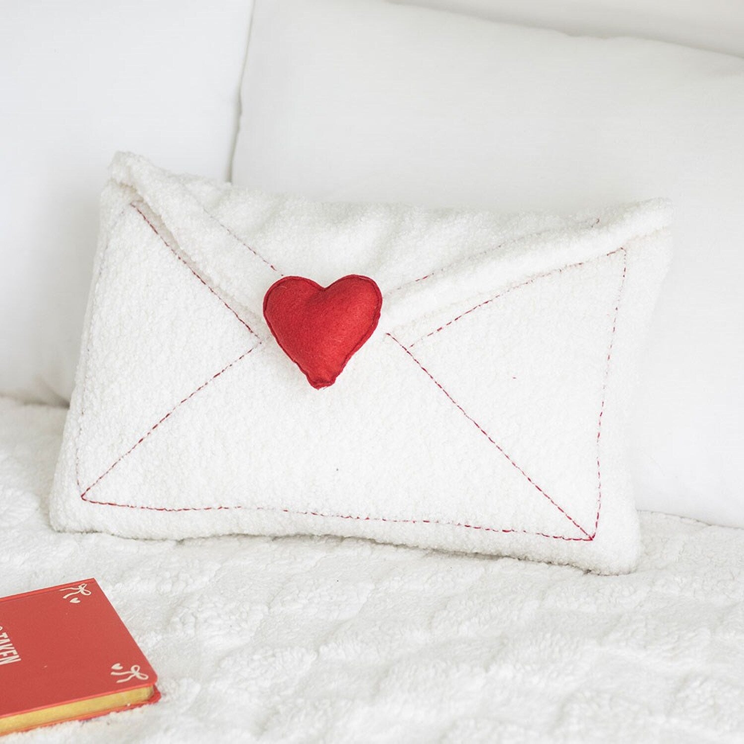Love Letter Embroidered Boucle Cushion on a white bed with a red book showing the envelope front with red heart detail