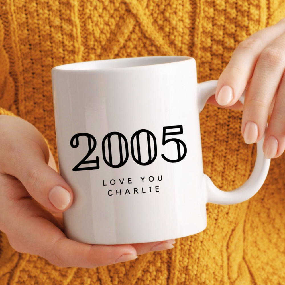 White mug with '2005 LOVE YOU CHARLIE' text held by a person against a yellow textured background