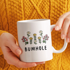 White mug with floral designs and 'BUMHOLE' text held by hands against a yellow textured background