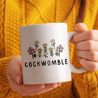 Person holding a white mug with floral design and 'COCKWOMBLE' text against a yellow textured background