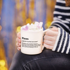 Person holding a mug with 'Pisces' definition text and colorful marshmallows against a blurred background.