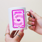 White mug with Aries zodiac design held by a hand against a beige background