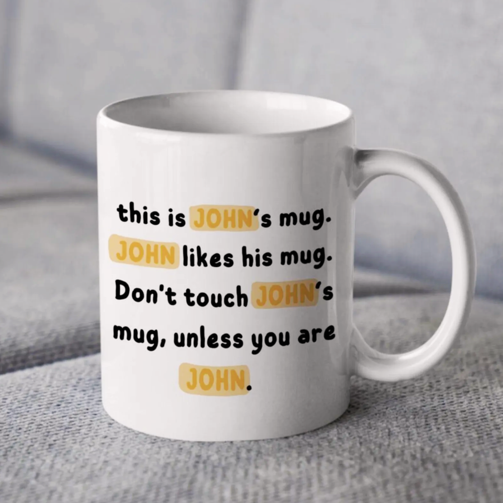 White mug with text 'this is JOHN's mug. JOHN likes his mug. Don't touch JOHN's mug, unless you are JOHN.' on a gray surface.