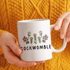 White mug with floral design and 'COCKWOMBLE' text held by a person against a yellow textured background