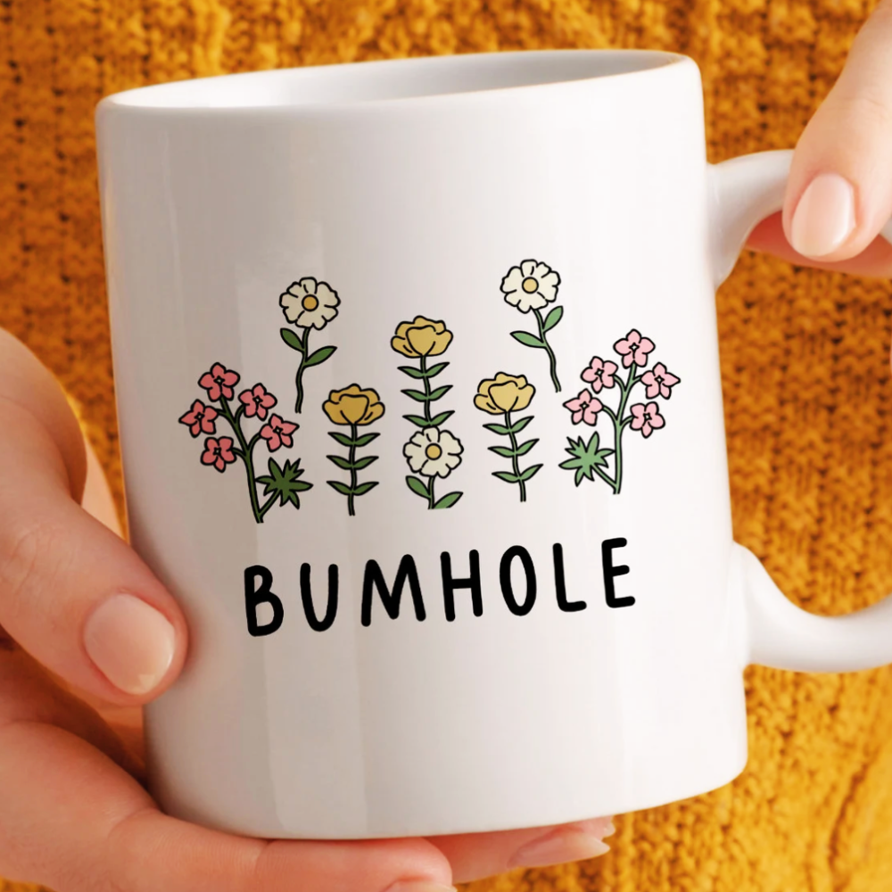 White mug with floral design and 'BUMHOLE' text held by a hand against an orange textured background