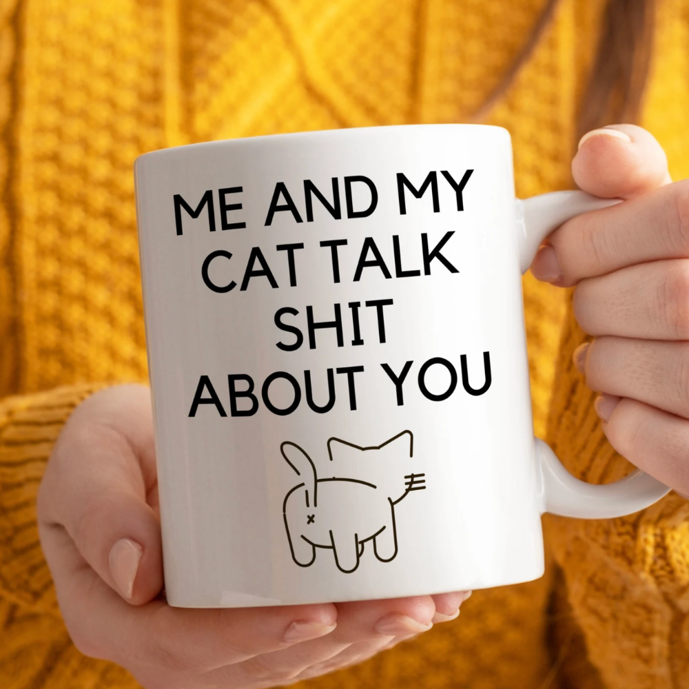 Person holding a mug with text 'Me and my cat talk shit about you' against a yellow textured background