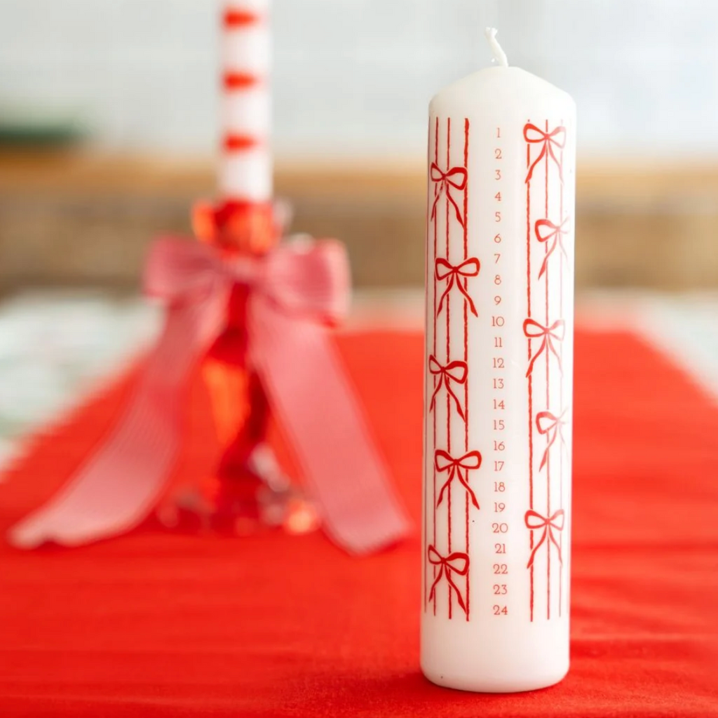 A white Advent candle with red printed numbers 1 through 24 and red bow motifs, standing upright on a red fabric surface. A second blurred candle with a pink ribbon is in the background.