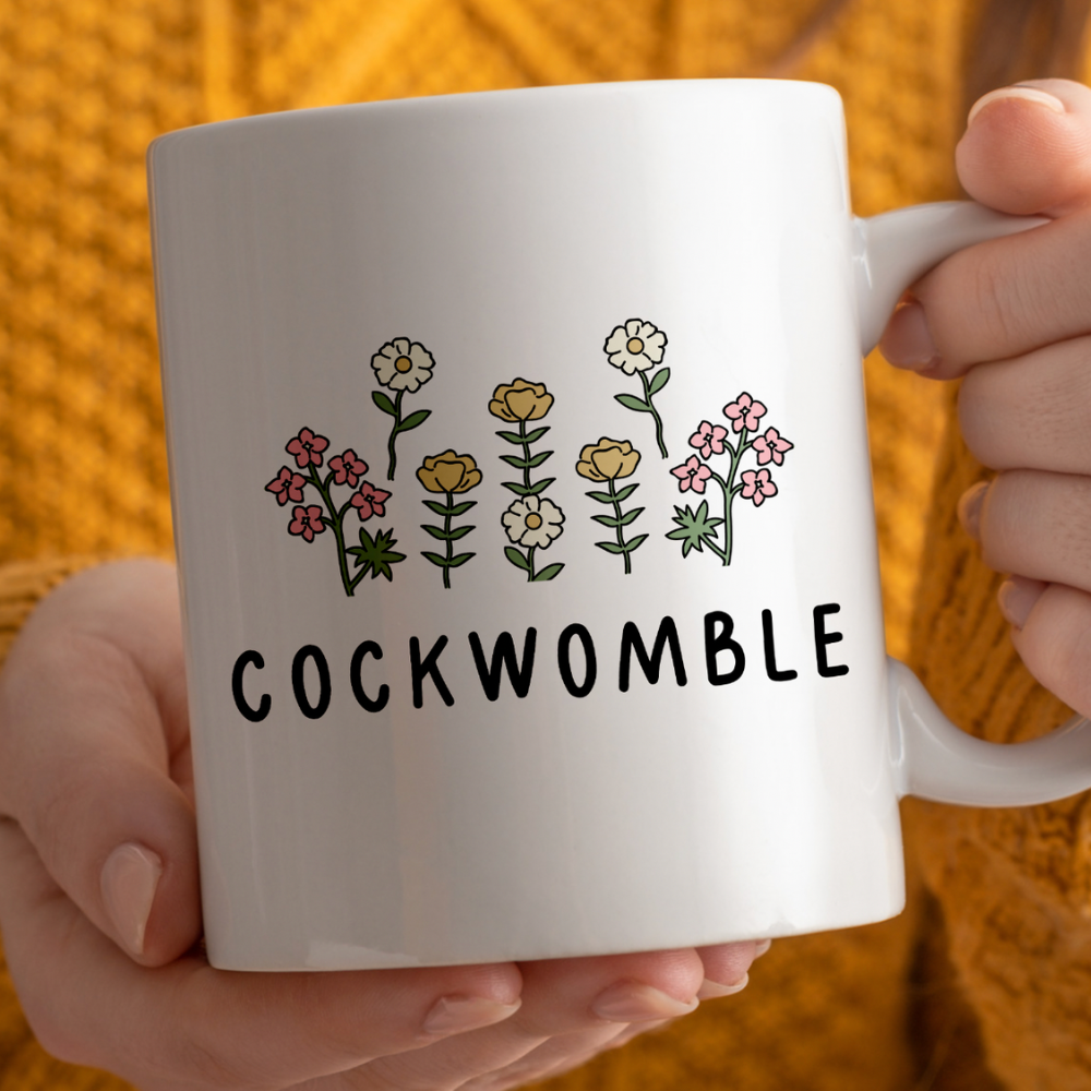 White mug with floral designs and 'COCKWOMBLE' text held by a person against an orange background