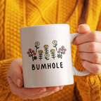Person holding a white mug with floral designs and the word 'BUMHOLE' against a yellow textured background.