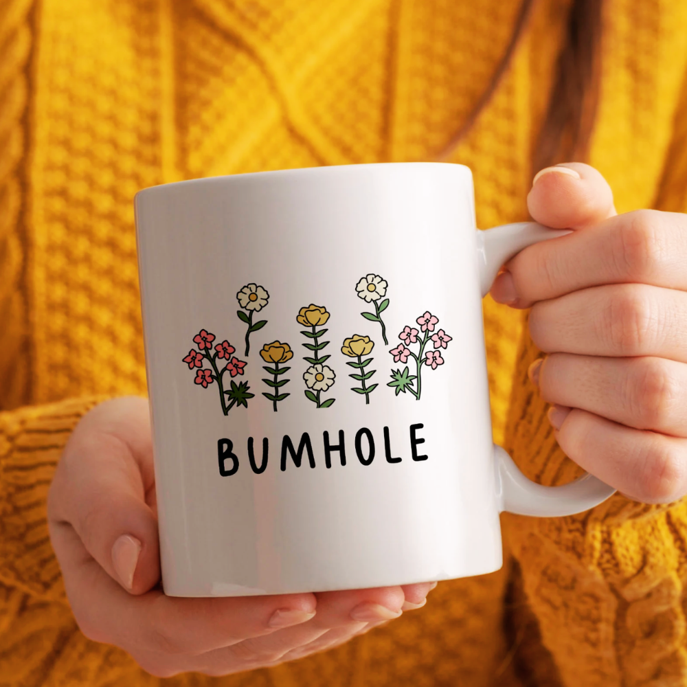 Person holding a white mug with floral designs and the word 'BUMHOLE' against a yellow textured background.