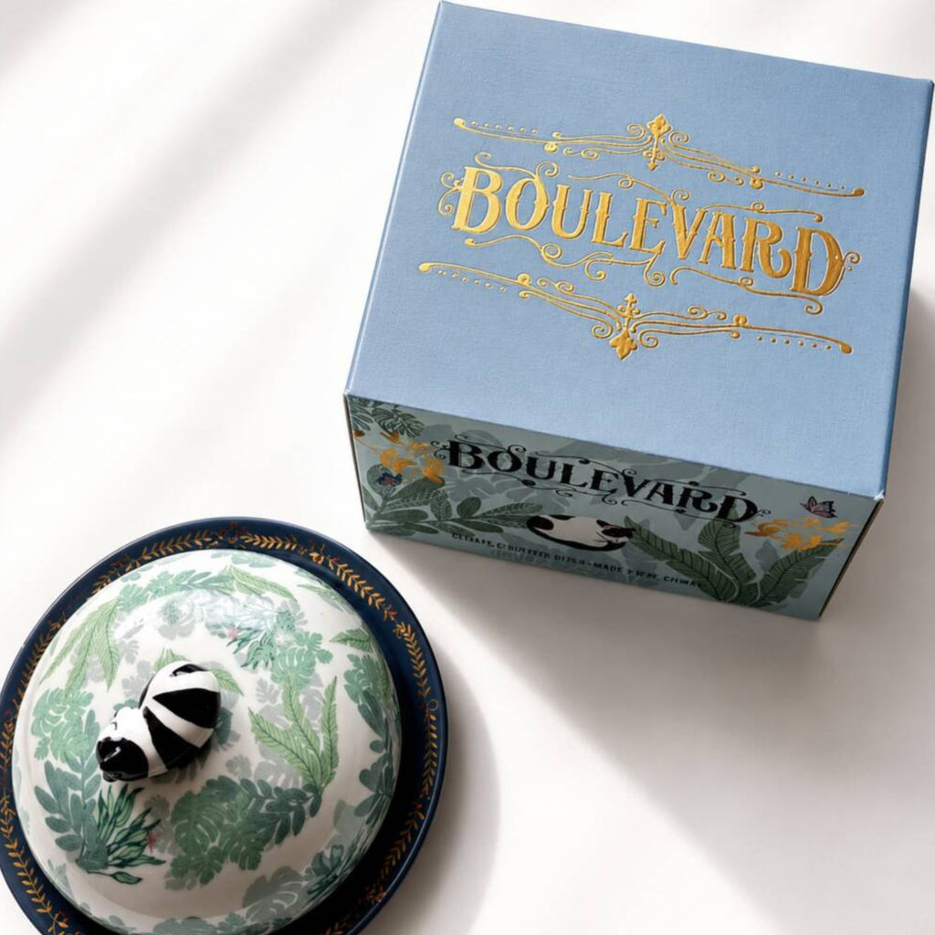 A light blue Boulevard gift box with gold lettering sits beside a matching ceramic dish with a lid. The dish has a white base with green botanical patterns and a black-and-white striped frog figurine on top.