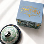 A light blue Boulevard gift box with gold lettering sits beside a matching ceramic dish with a lid. The dish has a white base with green botanical patterns and a black-and-white striped frog figurine on top.