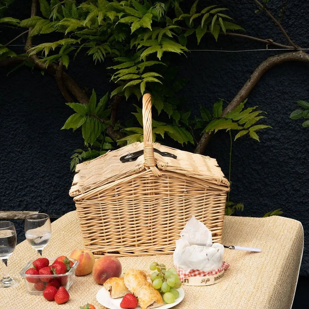 A wicker picnic basket with a house-shaped lid sits on a table outdoors, surrounded by food items including strawberries, grapes, bread, cheese, and wine glasses, with green foliage and a dark textured wall in the background.