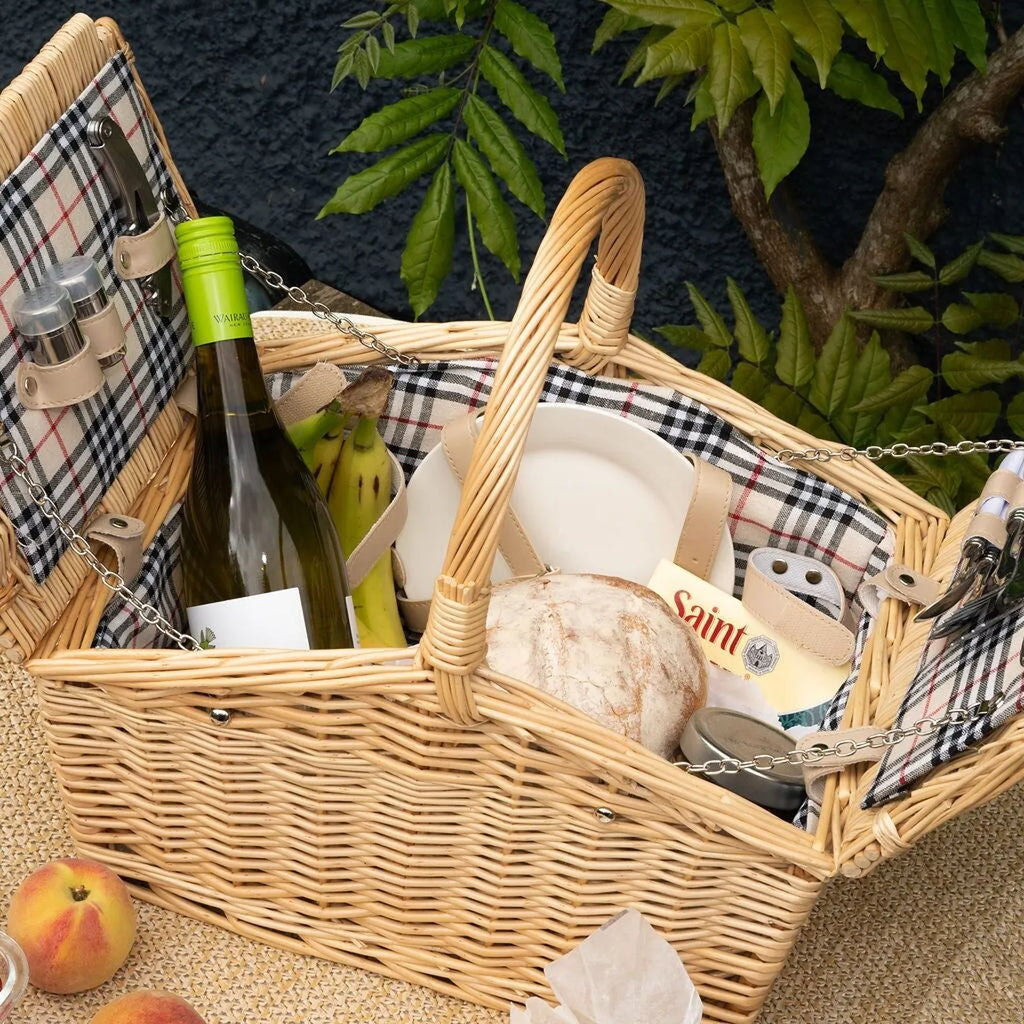 A wicker picnic basket filled with food items including a bottle of wine, bread, fruit, and cheese, set against a dark textured background with green foliage.