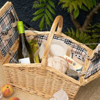 A wicker picnic basket filled with food items including a bottle of wine, bread, fruit, and cheese, set against a dark textured background with green foliage.