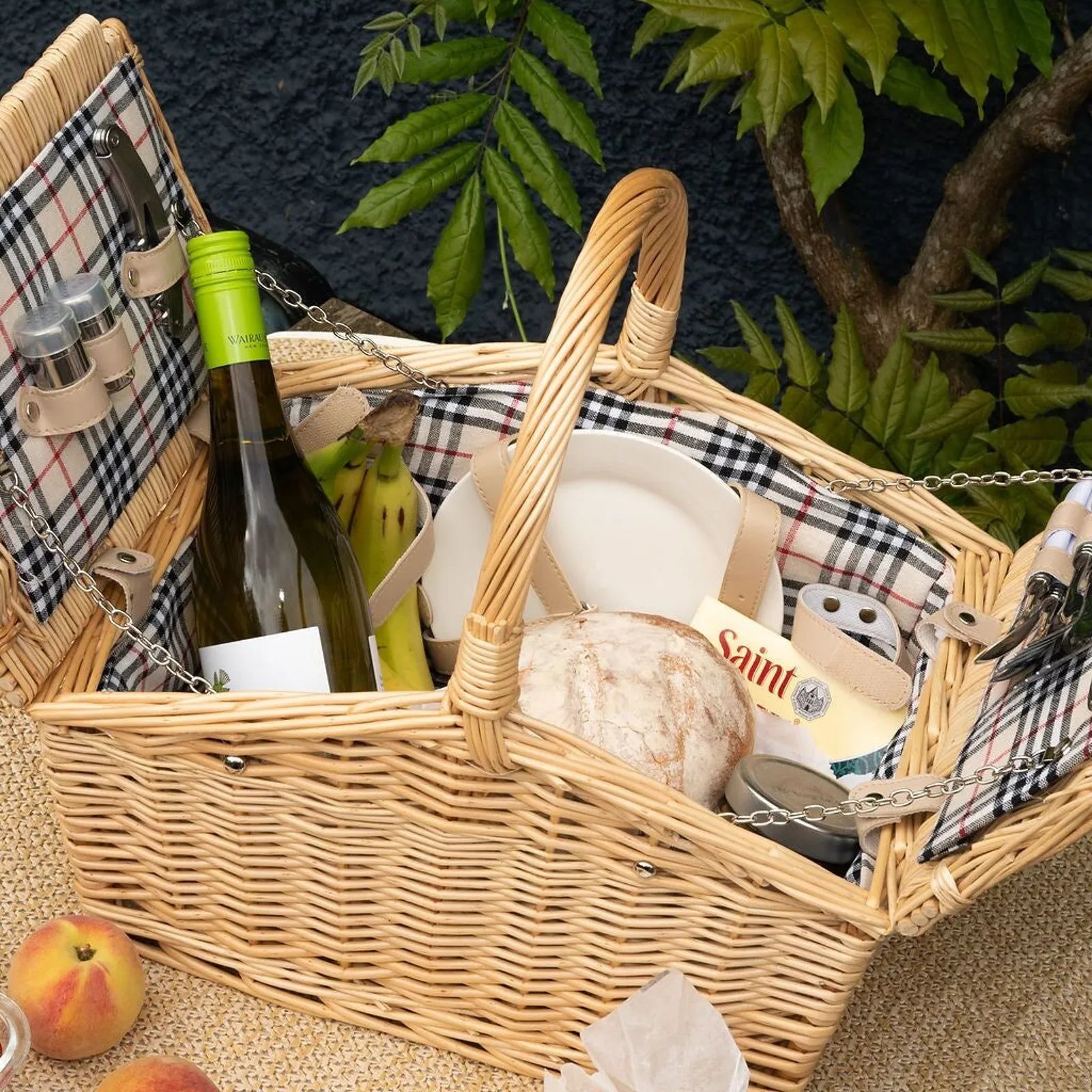 A wicker picnic basket filled with food items including a bottle of wine, bread, fruit, and cheese, set against a dark textured background with green foliage.