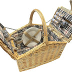 A classic wicker picnic basket with a beige and black plaid fabric lining, shown open to display its contents, including a white plate with a beige strap, metal canisters, and a picnic blanket on a white background.
