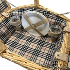 A wicker picnic basket with a beige and black plaid fabric lining, shown with a white plate and two clear wine glasses inside on a white background.