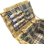 A wicker picnic basket with a beige, black, and red plaid fabric lining, shown open to display a matching cutlery set with white handles and a glass cup inside.