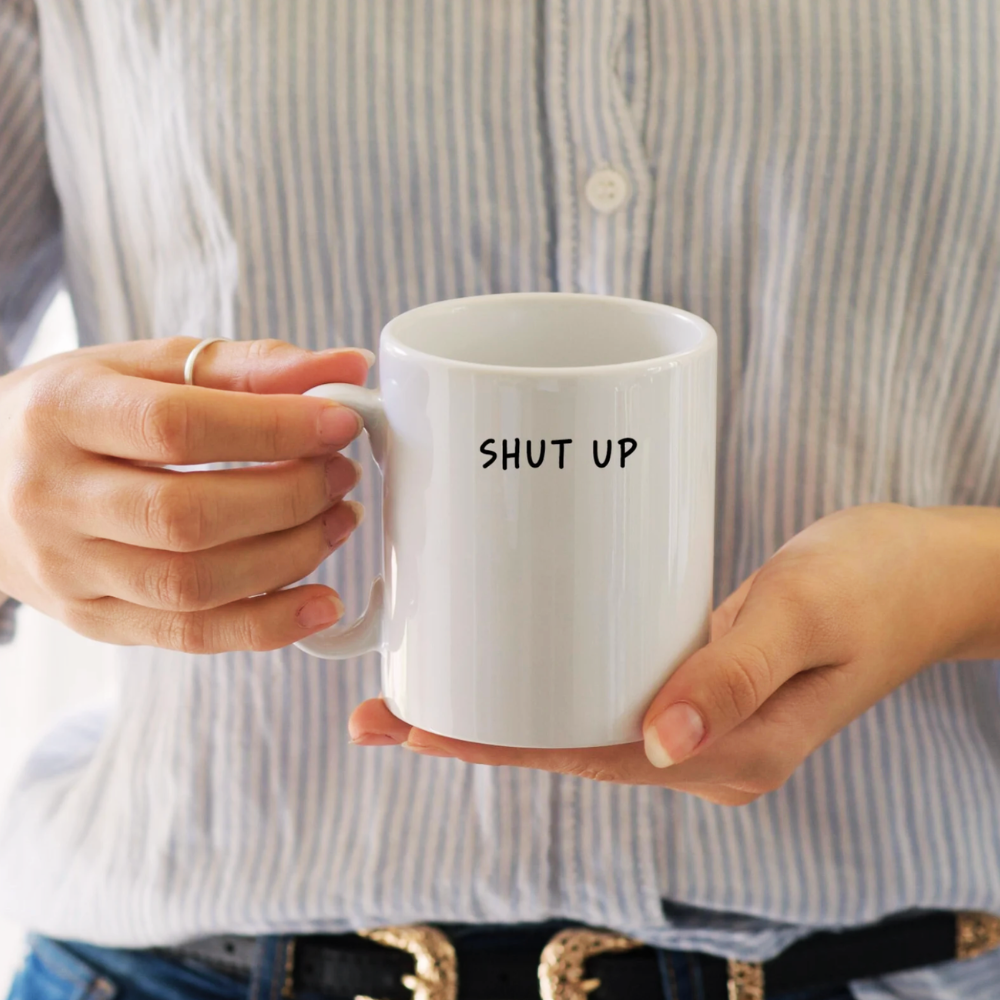 Person holding a white mug with 'SHUT UP' text, wearing a striped shirt.