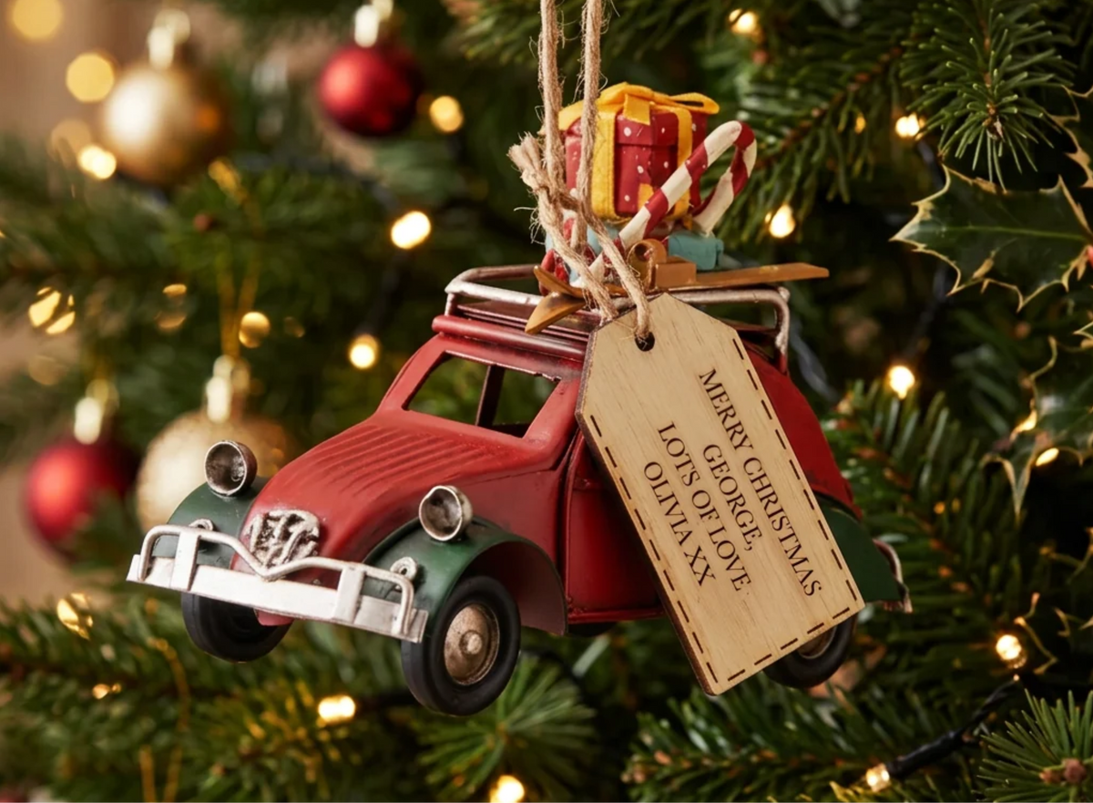 A red vintage toy car Christmas ornament hanging on a decorated Christmas tree, with a hanging tag that reads 'Merry Christmas from Colorado' and a small wrapped gift on top.