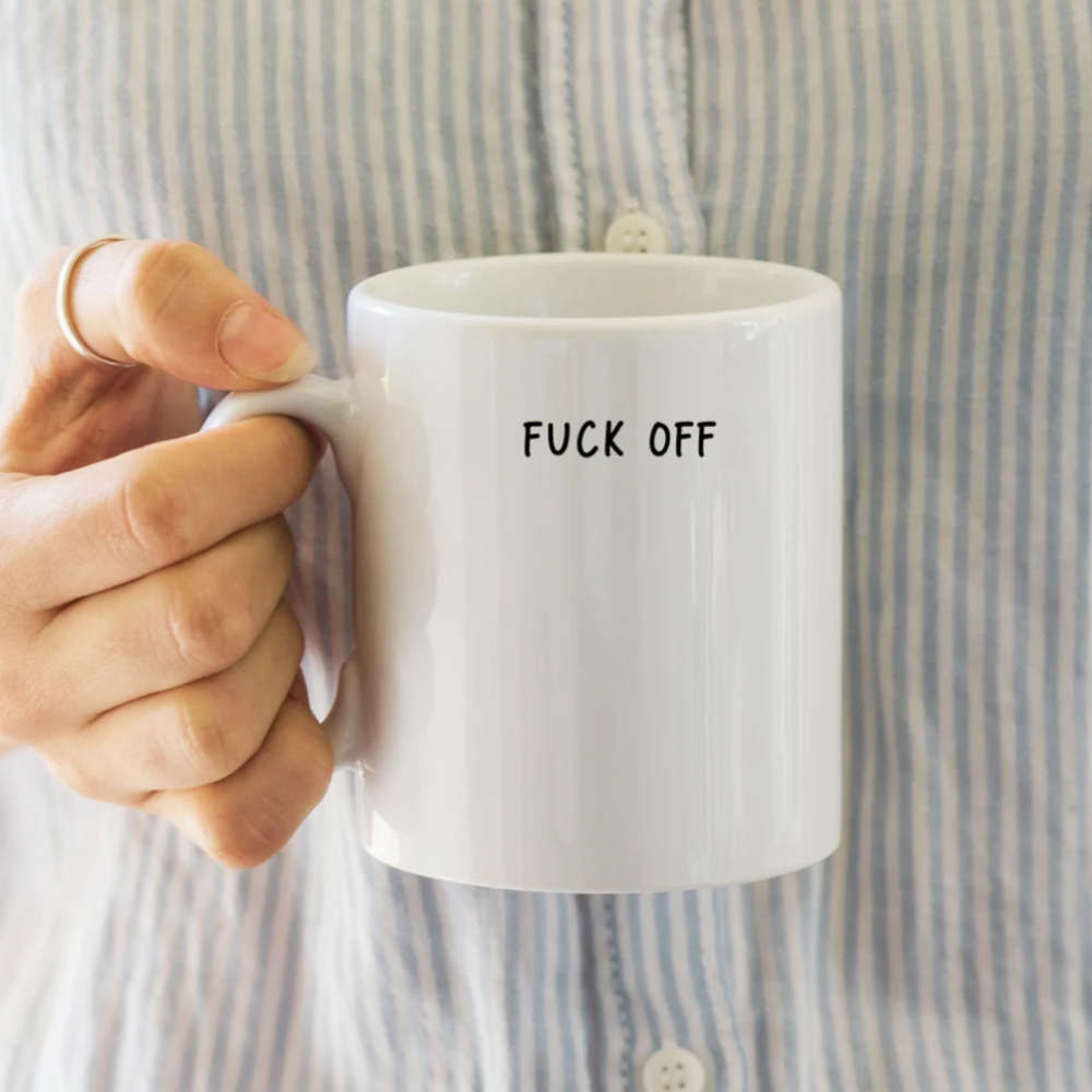 White mug with 'FUCK OFF' text held by a person wearing a striped shirt.