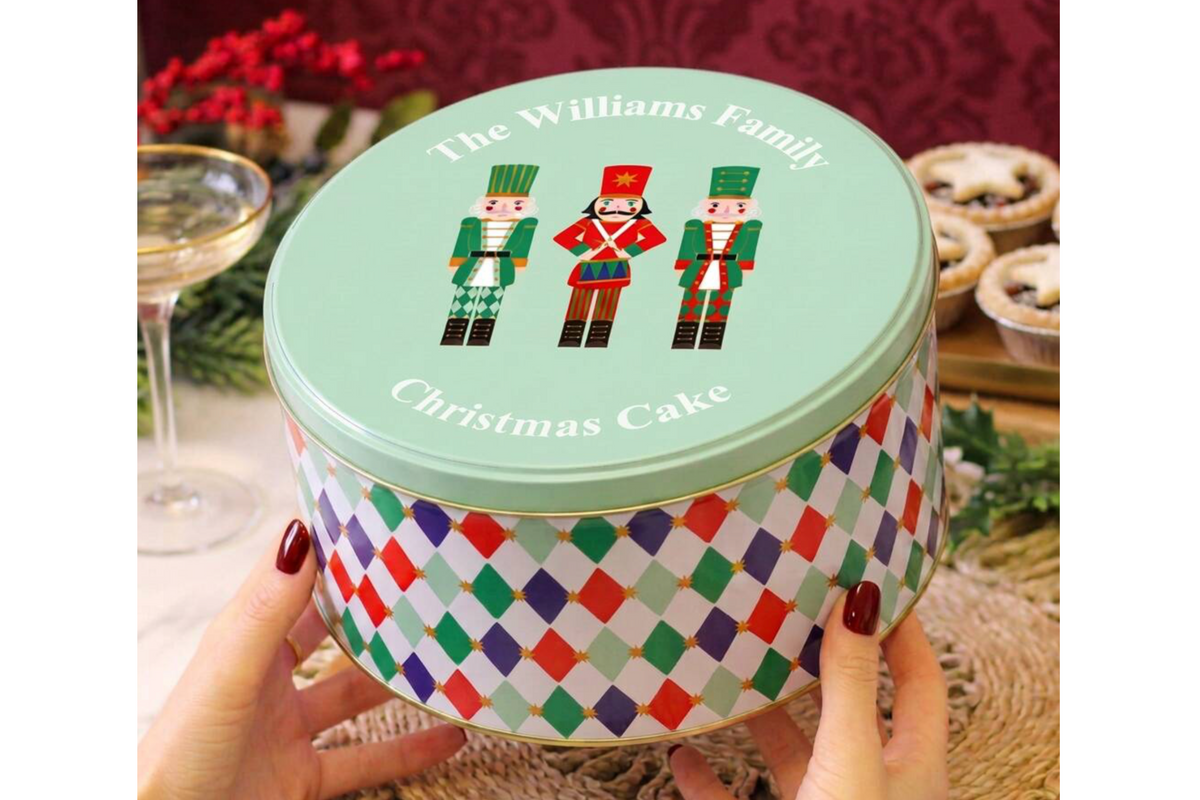 A round metal tin with a mint green lid decorated with three nutcracker figures, held by hands against a festive holiday background with a wine glass and pastries.