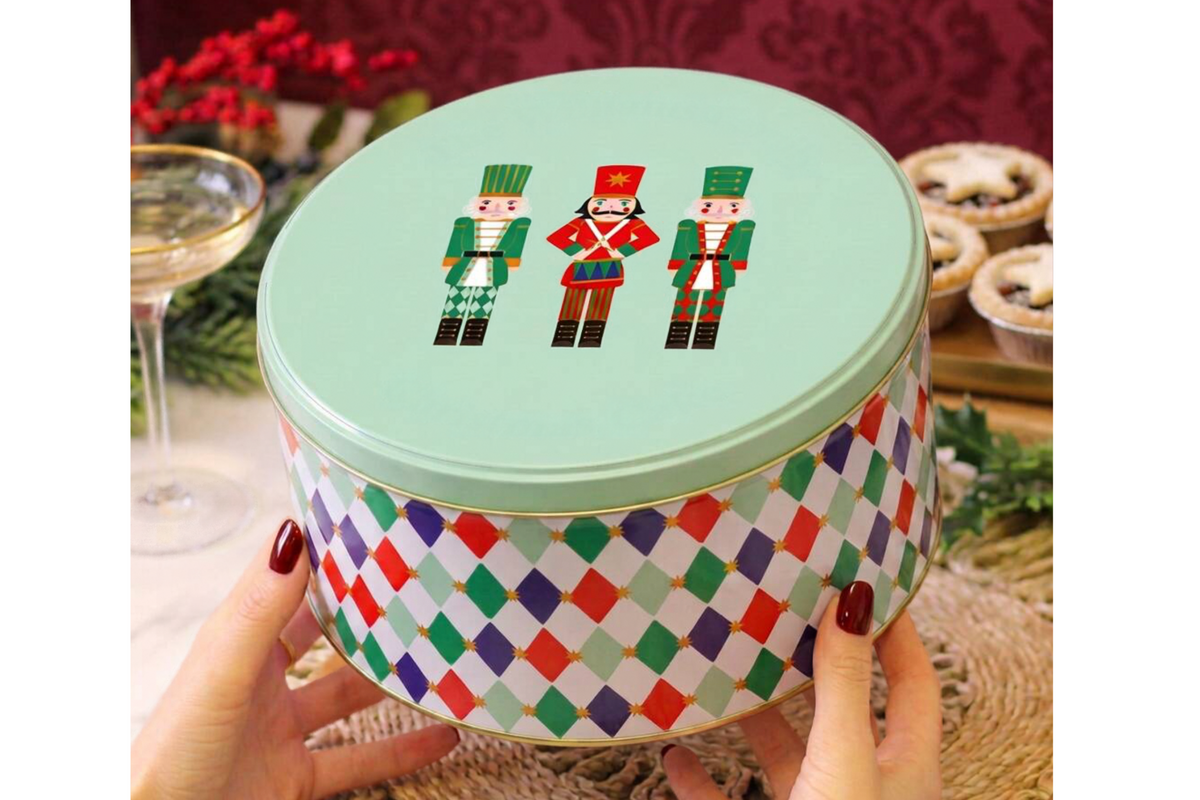 A round metal tin with a mint green lid decorated with three nutcracker figures, held by hands against a festive holiday background with a wine glass and pastries.