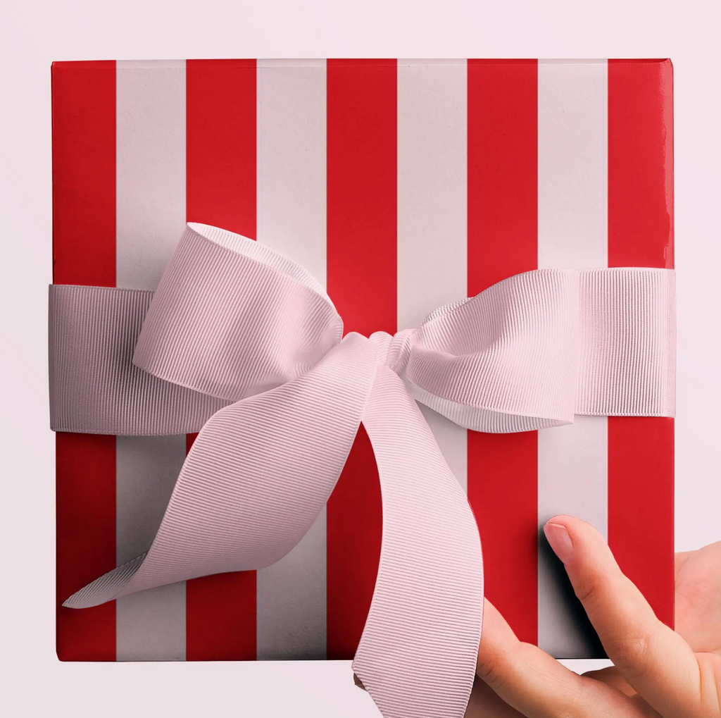A square gift box wrapped in red and white vertically striped paper, tied with a wide pale pink ribbon and bow, held from the right side by a person's hand against a light pink background.