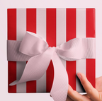A square gift box wrapped in red and white vertically striped paper, tied with a wide pale pink ribbon and bow, held from the right side by a person's hand against a light pink background.