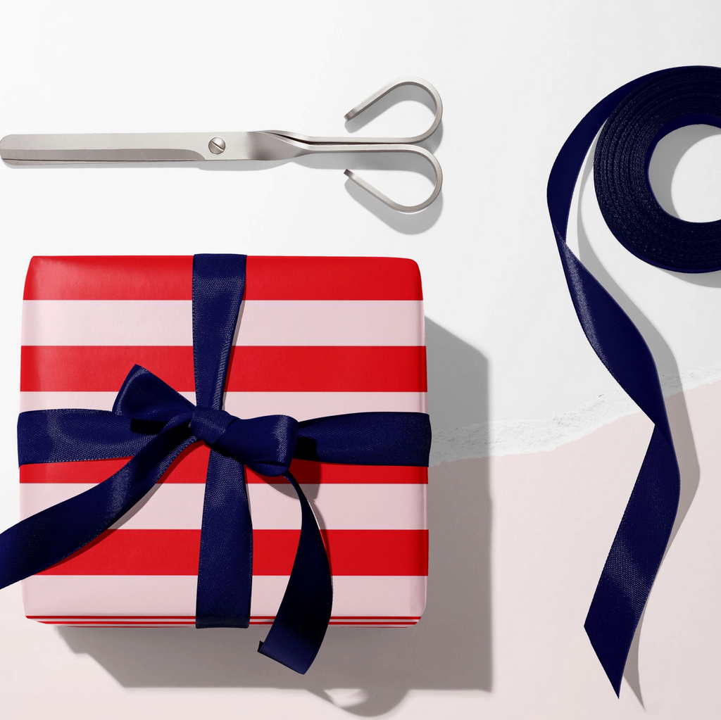 A wrapped gift box with red and white horizontal stripes and a navy blue ribbon bow, styled with silver scissors and a roll of matching ribbon on a white background.
