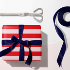 A wrapped gift box with red and white horizontal stripes and a navy blue ribbon bow, styled with silver scissors and a roll of matching ribbon on a white background.