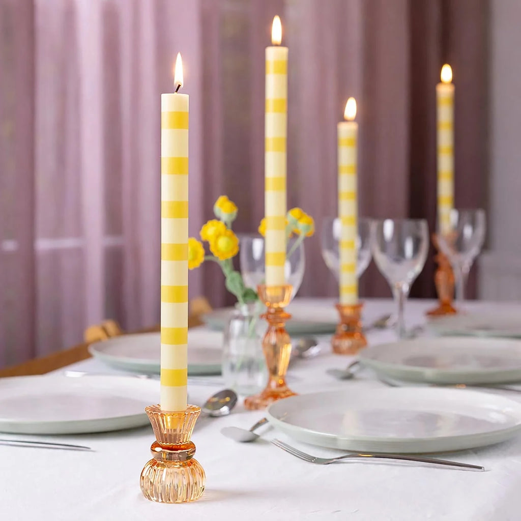 Lemon Stripe Dinner Candle lit in an amber glass candleholder on a white dinner table with glassware and yellow flowers