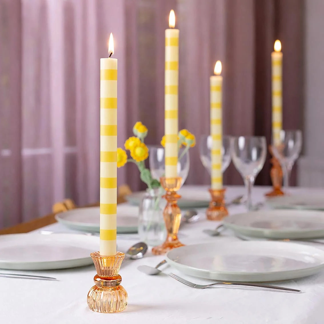 Lemon Stripe Dinner Candle lit in an amber glass candleholder on a white dinner table with glassware and yellow flowers