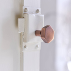 A close-up of a hexagonal copper door latch bolt and strike plate set against a white painted wooden door.