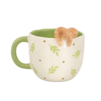 A ceramic mug with a cream-colored exterior decorated with green leaves and brown dots, a light green handle and interior, and a small 3D sloth figurine resting on the rim.