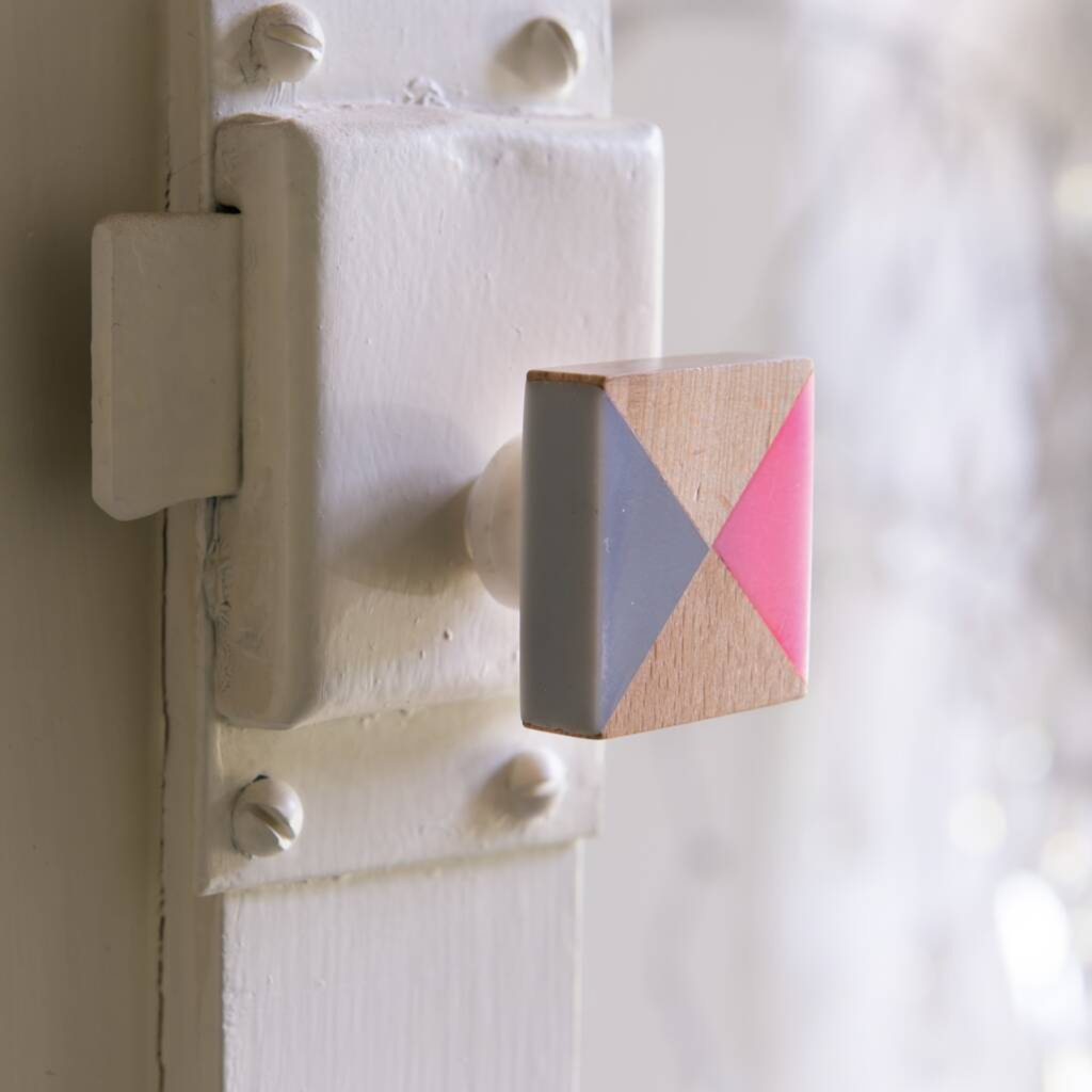 A close-up of a square wooden door knob mounted on a white painted door latch. The knob features a geometric color-blocked design with pink, gray, and natural wood triangles.