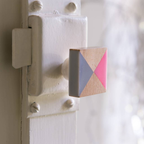 A close-up of a square wooden door knob mounted on a white painted door latch. The knob features a geometric color-blocked design with pink, gray, and natural wood triangles.