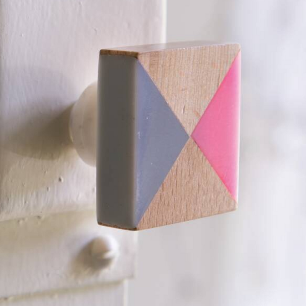 A close-up of a square wooden cabinet knob with a geometric design of pink, gray, and natural wood triangles, mounted on a white painted door.