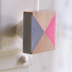 A close-up of a square wooden cabinet knob with a geometric design of pink, gray, and natural wood triangles, mounted on a white painted door.
