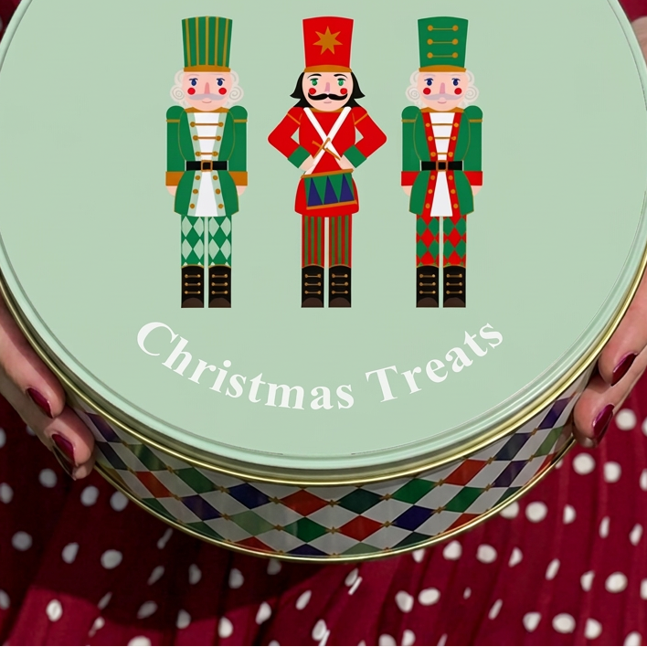 A round tin of Christmas treats held by a person wearing a red and white polka dot dress. The lid is pale mint green with white text reading 'Rowan & Hallies Christmas Treats' and an illustration of three nutcracker figures in green, red, and black.