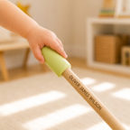 A close-up of a child's hand holding a personalized wooden stick with a light green handle. The wooden shaft is engraved with the name 'OLIVER JAMES WILSON' and is set against a softly blurred indoor room background.