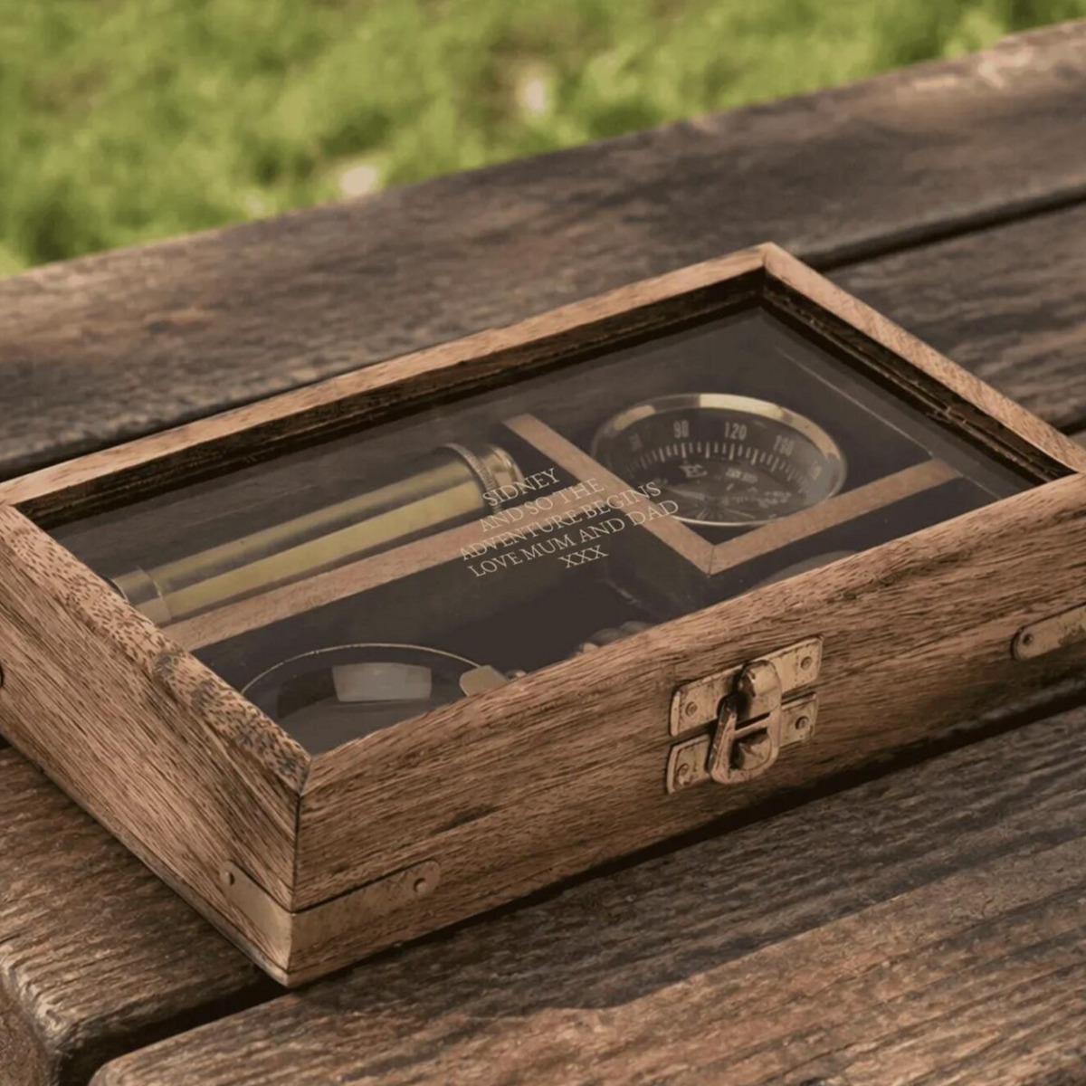 A rectangular wooden presentation box with a clear glass top lid, shown resting on a rustic wooden table outdoors. Inside the box is a vintage-style pocket watch with a black dial and gold-toned details.
